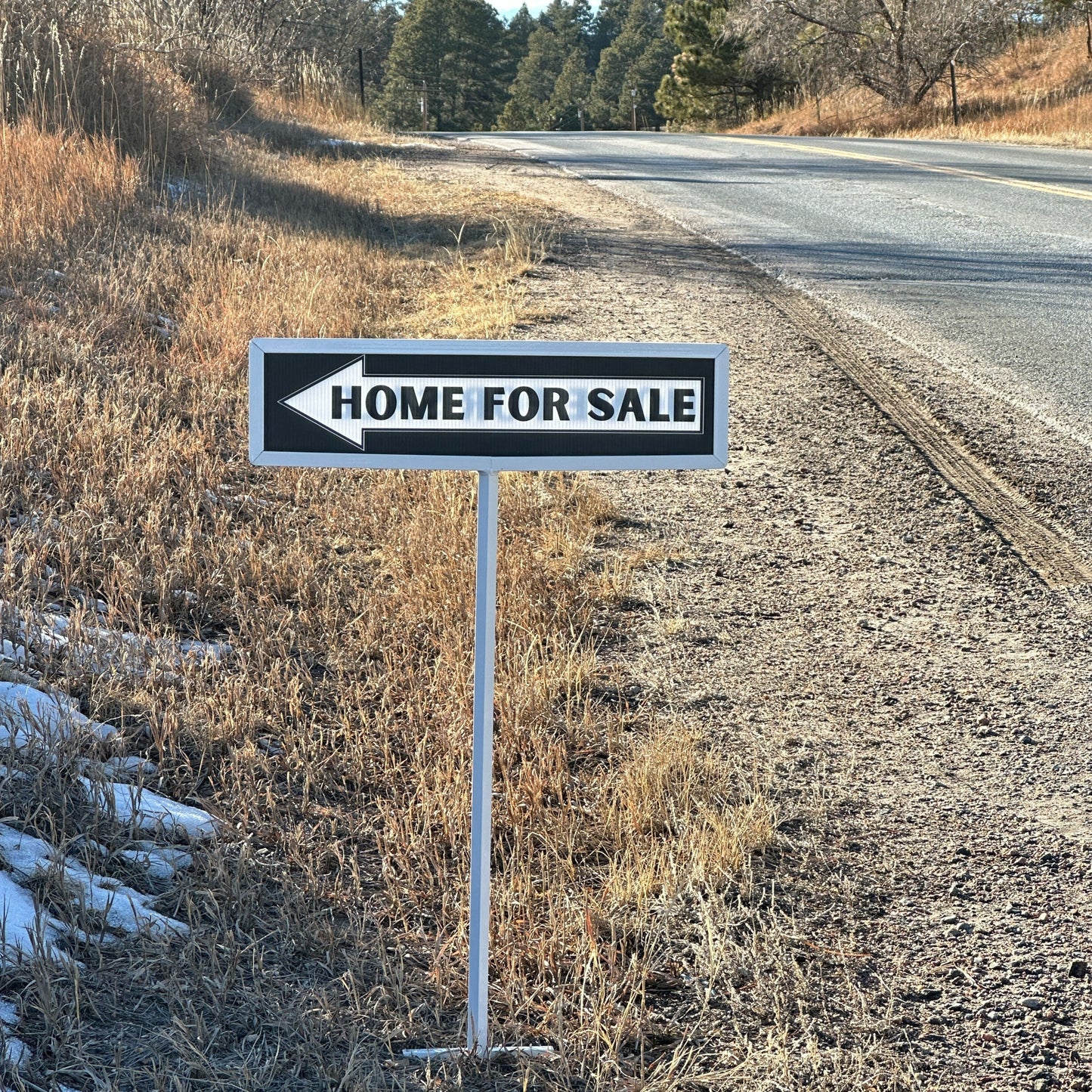 Home for sale sign on the side of a rural road with trees in the background.