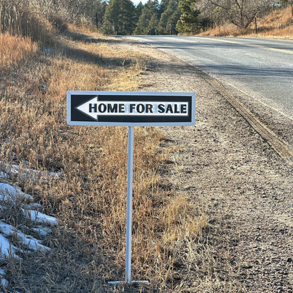 Home for sale sign on the side of a rural road with trees in the background.