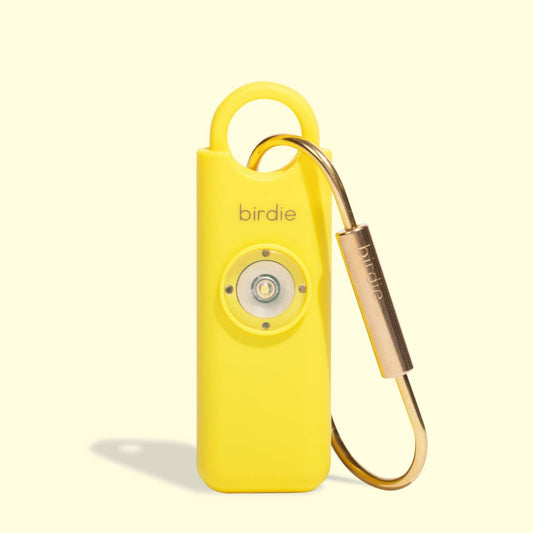A yellow personal safety alarm with a solid brass keychain and a flashing strobe-light feature, branded as She's Birdie.